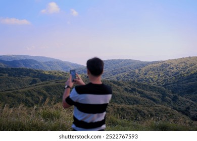 Man in black and white stripes, back facing holding phone, admiring mountain view with green hills in sunlight under blue cloudy sky. Nature, landscape, outdoor adventure, peaceful, serene, mountains. - Powered by Shutterstock