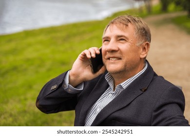 Man in a blazer enjoying a friendly phone call while sitting by a serene lakeside park during a sunny afternoon in a peaceful outdoor setting - Powered by Shutterstock