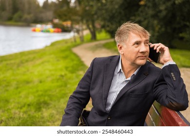 Man in a blazer enjoying a friendly phone call while sitting by a serene lakeside park during a sunny afternoon in a peaceful outdoor setting. High quality photo - Powered by Shutterstock