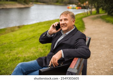 Man in a blazer enjoying a friendly phone call while sitting by a serene lakeside park during a sunny afternoon in a peaceful outdoor setting - Powered by Shutterstock