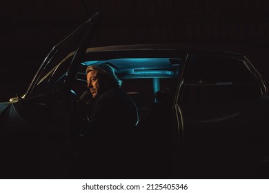 Man With Blond Hair Sits Inside An American Classic Muscle Car At Night.