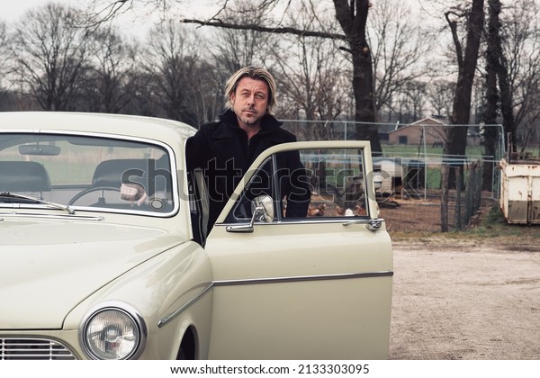 Man with blond hair and stubble in a\
black coat stands by a classic car in the\
countryside.