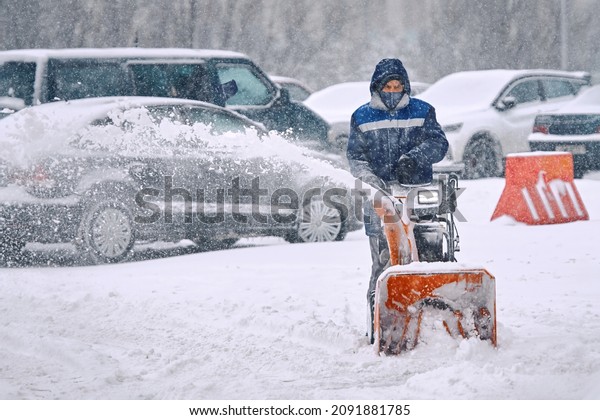 Man\
blow snow from parking lot with snow blower machine during\
snowfall. Worker blowing snow during blizzard. Man using\
snowblower, clearing snow from road at parking\
lot