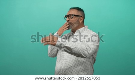 Man blowing a kiss with both hands to camera in teal studio, wearing glasses and white dotted shirt; affection playful greeting.