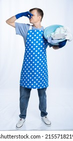 Man In Blue Apron, Rubber Gloves, Blue T-shirt, Jeans. Homeowner Holding A Basin With Dirty Laundry Pinched His Nose With His Hand. White Background.