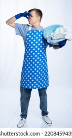 Man In Blue Apron, Rubber Gloves, Blue T-shirt, Jeans. Homeowner Holding A Basin With Dirty Laundry Pinched His Nose With His Hand. White Background.