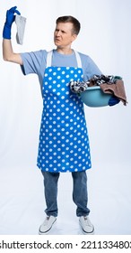 Man In Blue Apron, Rubber Gloves, Blue T-shirt, Jeans. The Homeowner Holds A Basin With Dirty Laundry And A Sock. White Background.