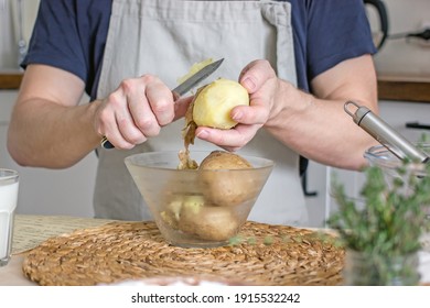 A Man In A Blue Apron, Without A Face, Prepares A Potato Dish In His Kitchen, Holds A Potato In His Hands And Peels It, Authentic Home Cooking. Male Home Cook Hobby