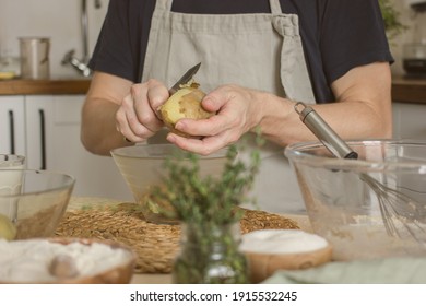 A Man In A Blue Apron, Without A Face, Prepares A Potato Dish In His Kitchen, Holds A Potato In His Hands And Peels It, Authentic Home Cooking. Male Home Cook Hobby