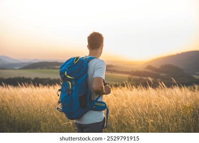 Man with a blue backpack stands in a grassy field, gazing at a breathtaking sunset over rolling hills. The scene captures the tranquility of nature, adventure, and personal reflection as the sun sets. - Powered by Shutterstock