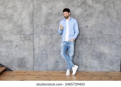 A man in a blue button-down shirt and jeans stands with one leg crossed against a concrete wall, looking at his smartphone with a slight smile. The man has a beard and short dark hair, copy space - Powered by Shutterstock