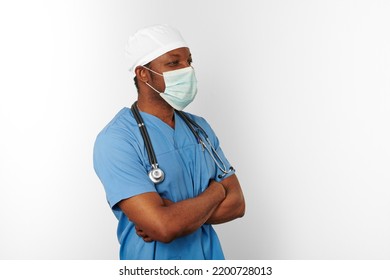 Man In Blue Coat White Cap And Surgeon Mask With Crossed Arms Isolated On White Background. Focused Adult African American Practicing Surgeon Portrait, Confident Look