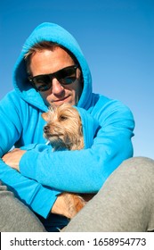 Man In Blue Hoody Sitting Outdoors Cuddling Small Dog In Matching Outfit