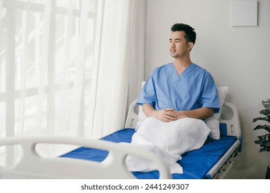 A man in a blue hospital gown sits on a hospital bed with a white blanket. He looks tired and is staring out the window - Powered by Shutterstock