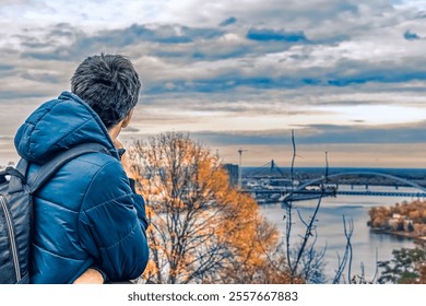 A man in a blue jacket with a backpack looks out over the scenic view of Kyiv, Ukraine, with the Dnipro River and a bridge visible in the distance under a cloudy sky - Powered by Shutterstock