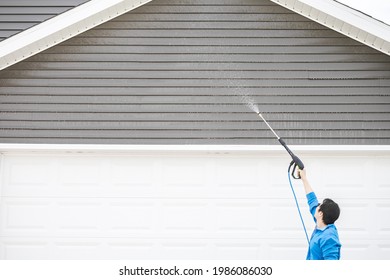 Man In Blue Jacket Clean Dusk And Dirt From Exterior Siding Under The Roof At Front Door Garage By High Pressure Nozzle Spray With Water Soap Cleaner, Weekend Chore. Washer Machine Of Cleansing House.