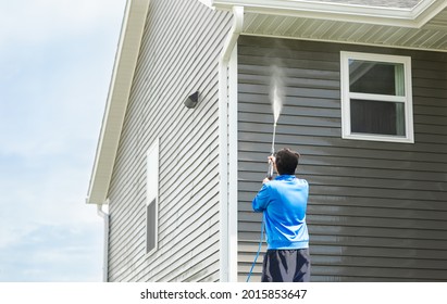 Man in blue jacket cleans dusk and dirt from exterior siding and under roof with a high-pressure nozzle spray with water soap cleaner. Wash a house during the day. Home maintenance service concept.