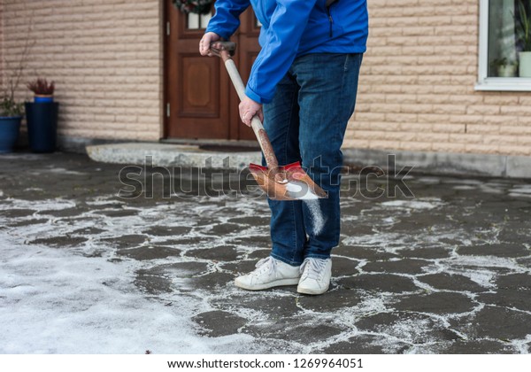 A man in\
blue jacket, jeans and white sneakers is using a shovel to sprinkle\
road salt on the paving slabs in front of a house to remove the ice\
and prevent a slipping\
accident.