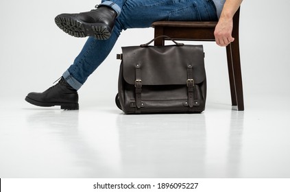 Man in a blue jeans and black boots sits on a chair with a brown men's shoulder leather bag for a documents and laptop on a white floor. Mens leather satchel, messenger bags, handmade briefcase. - Powered by Shutterstock