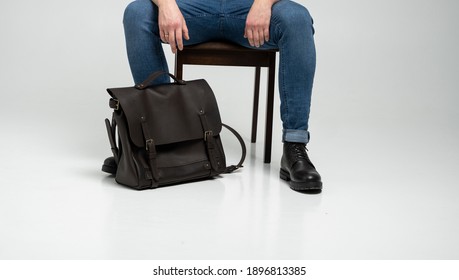 Man In A Blue Jeans And Black Boots Sits On A Chair With A Brown Men's Shoulder Leather Bag For A Documents And Laptop On A White Floor. Mens Leather Satchel, Messenger Bags, Handmade Briefcase.