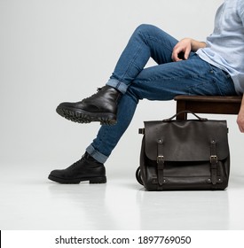 Man In A Blue Jeans And Black Boots Sits On A Chair With A Brown Men's Shoulder Leather Bag For A Documents And Laptop On A White Floor. Mens Leather Satchel, Messenger Bags, Handmade Briefcase.