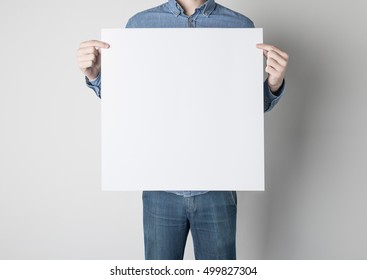 Man In Blue Jeans And Shirt Is Standing In Front Of The White Wall While Holding A Square White Paper Poster.