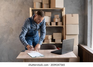A  man in a blue overalls prepares goods for shipment, makes notes with a pen. Online sales with start-up and small business, conceptual work from home and online shopping with e-commerce business