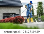 A man in blue overalls and yellow boots cleans a driveway with a pressure washer, surrounded by lush greenery and vibrant flowers on a bright afternoon.