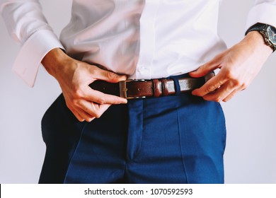 A man in blue pants and a white shirt buttoned a brown leather trouser belt. He has a watch on his hand