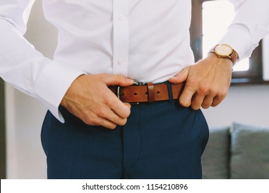 A man in blue pants and a white shirt buttoned a brown leather trouser belt. He has a watch on his hand.