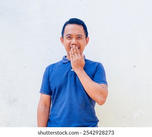 A man in a blue polo shirt stands against a plain background, covering his mouth with a smiling expression. - Powered by Shutterstock