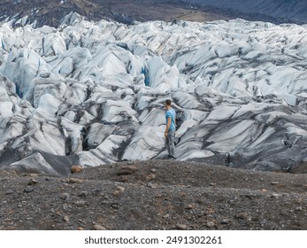 A man in a blue shirt and black pants stands on a rocky outcropping, gazing at a snow-covered glacier with ridges and valleys in Iceland. - Powered by Shutterstock