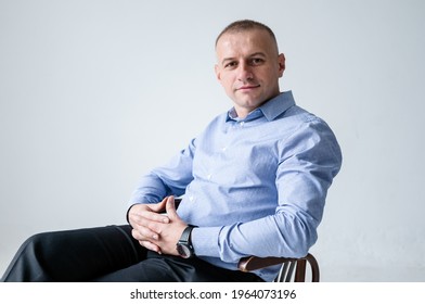 A Man In A Blue Shirt And Black Trousers Sits On A Black Stool On A White Background