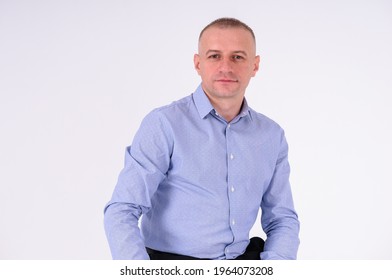 A Man In A Blue Shirt And Black Trousers Sits On A Black Stool On A White Background