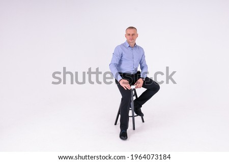 A man in a blue shirt and black trousers sits on a black stool on a white background