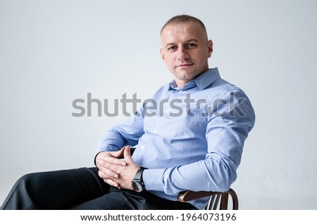 A man in a blue shirt and black trousers sits on a black stool on a white background