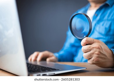 A man in a blue shirt holds a magnifying glass while using a laptop, symbolizing online research, investigation, cybersecurity, data analysis, or scrutiny in digital environments on a wooden desk.