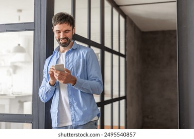 A man in a blue shirt is standing in an office hallway, leaning against a wall. He is looking down at his phone and smiling, copy space - Powered by Shutterstock