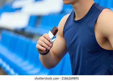 Man In A Blue Sports Jersey Rests In The Stand For Fans, Eating A Chocolate Bar. A Muscular Athlete Snacks On A Protein Bar While Resting After A Workout At The Stadium