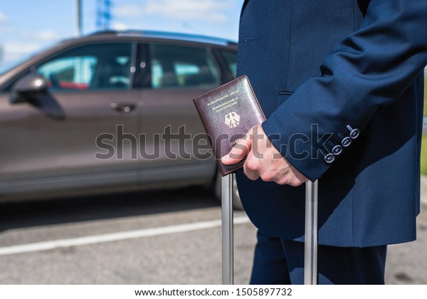 Man in a blue suit with a suitcase and germany\
passport at the airport parking on the backdrop of his car.\
Business trip concept