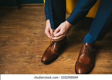 A man in a blue suit ties up shoelaces on brown leather shoes brogues on a wooden parquet background