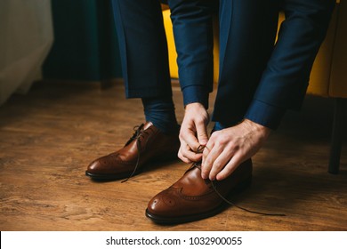 A man in a blue suit ties up shoelaces on brown leather shoes brogues on a wooden parquet background - Powered by Shutterstock