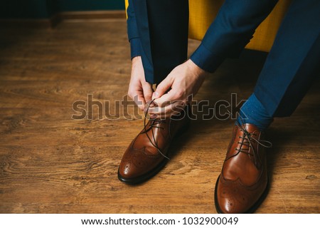 A man in a blue suit ties up shoelaces on brown leather shoes brogues on a wooden parquet background