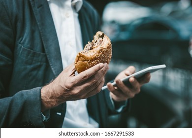 Man In A Blue Sute Working Online On His Smartphone While Lunchbreak Eating A Hamburger Or Sandwich Standing Outside Of Business Center.