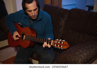 A man in a blue sweater plays an acoustic guitar on a brown sofa, emphasizing music and relaxation indoors. The cozy living room setting adds to the evening's peaceful atmosphere. - Powered by Shutterstock