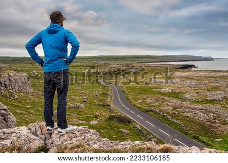 Man in blue travel jacket and hat standing on a edge of a mountain, stunning rough nature scenery with small road and tough terrain in the background. Burren, Ireland. Travel and tourism concept.