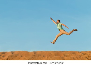man in a blue t-shirt and beige trousers jumps over crests of dunes in the desert under the heated sun high. The right hand is highly raised to the sun up.  orange sandy dunes and the clear blue sky