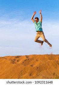 man in a blue t-shirt and beige trousers jumps over crests of dunes in the desert under the heated sun high. The right hand is highly raised to the sun up. orange sandy dunes and the clear blue sky