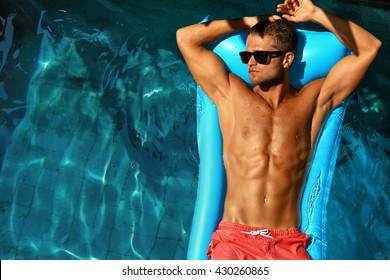 Man Body In Summer. Male In Pool. Beautiful Sexy Model In Fashion Sunglasses Tanning In Swimming Pool Water At Resort. Fit Guy With Healthy Sun Tan Skin Relaxing On Holidays Vacation. Summertime Relax