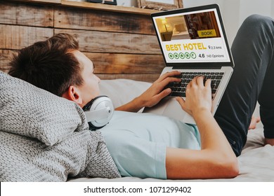 Man booking the best hotel for vacation by internet with a laptop computer. Hotel reservation website appears in the notebook screen while he types on the keyboard, lying and resting on the bed.